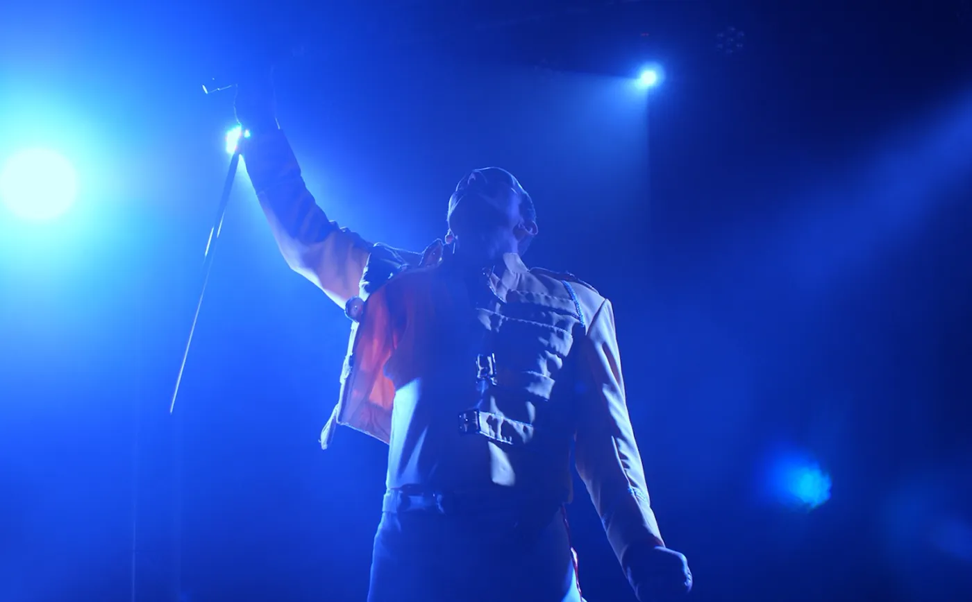 Eddie Devlin as Freddie Mercury in the yellow jacket, mic stand raised in a blue spotlight